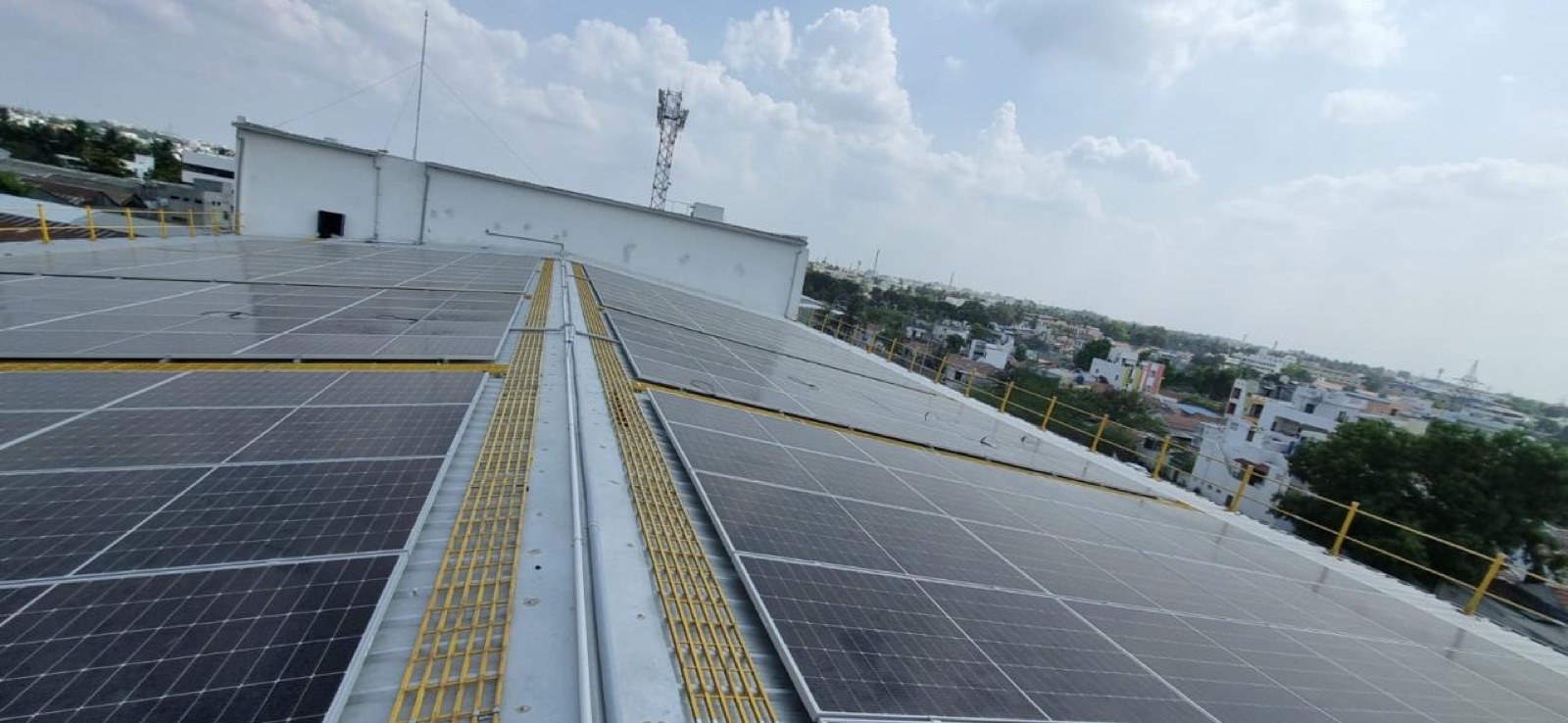 Wide industrial rooftop solar array with maintenance walkways, a telecom tower and town skyline on the horizon.